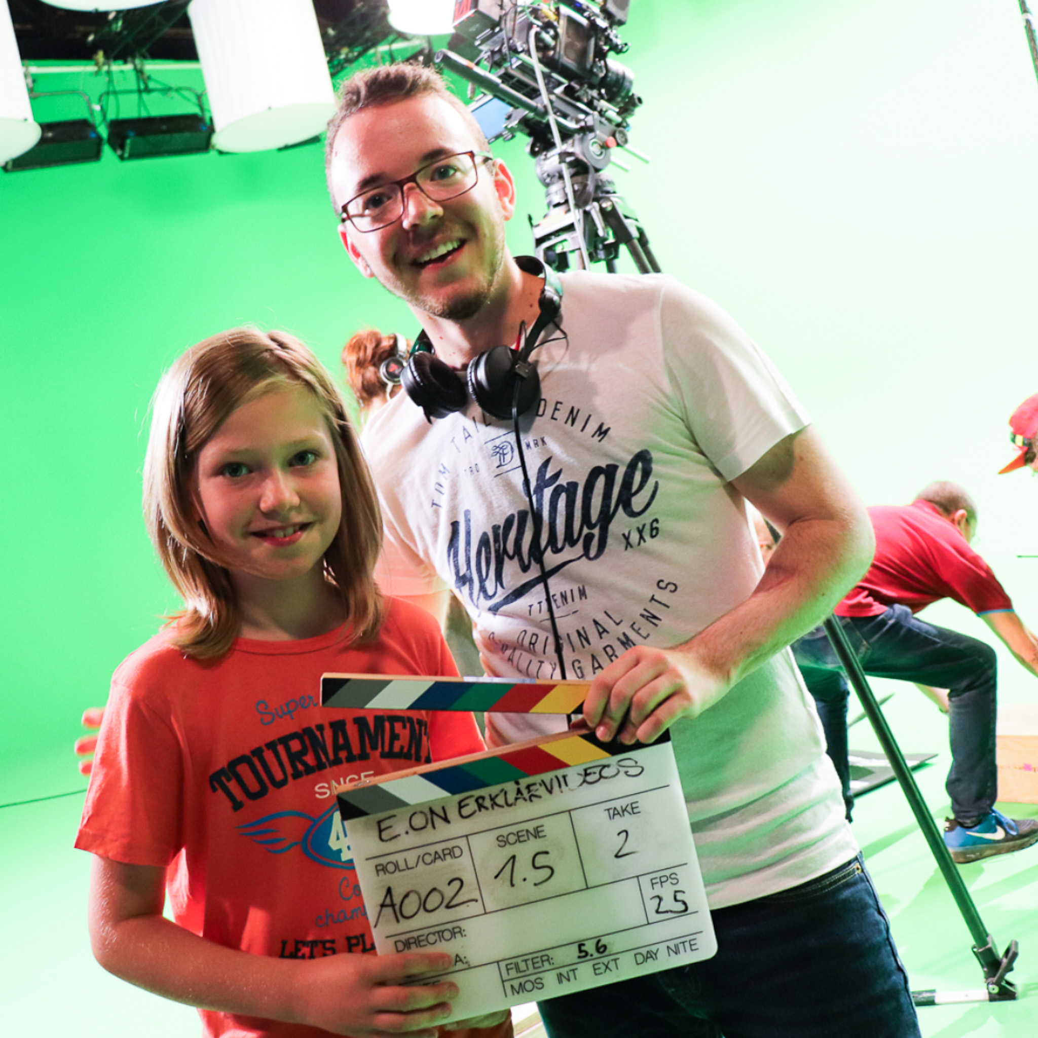 Erklärfilme Ein Kind steht neben einem Erwachsenen. Beide halten eine Filmklappe in der Hand. Der Erwachsene trägt zudem Kopfhörer um sein Hals