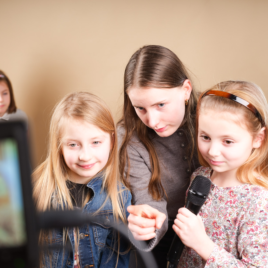 Eine Frau steht zwischen zwei Mädchen und zeigt mit ihrem Finger auf etwas. Das eine Mädchen hält dabei ein Mikrofon in einer Hand und das andere Mädchen trägt eine blaue Jeans Jacke.
