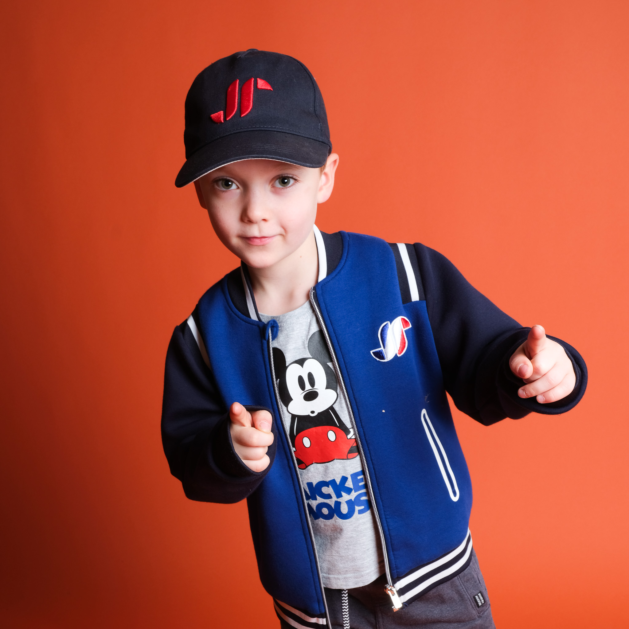 Kleiner Junge vor einem Orangen Hintergrund mit eine Capi und Jacke. Zeigt dabei auf die Kamera